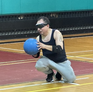 A resident kneels on the floor of a gym at the Chicago Park District holding a blue ball while playing goal ball.