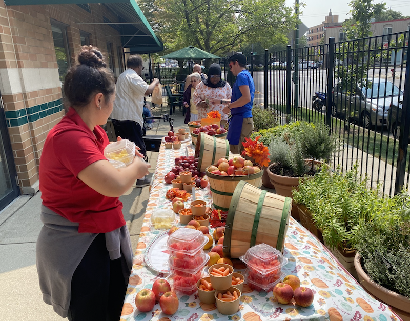 A group of people stand at tables with fresh apples outside of the Friedman Place building for a farmer's market.