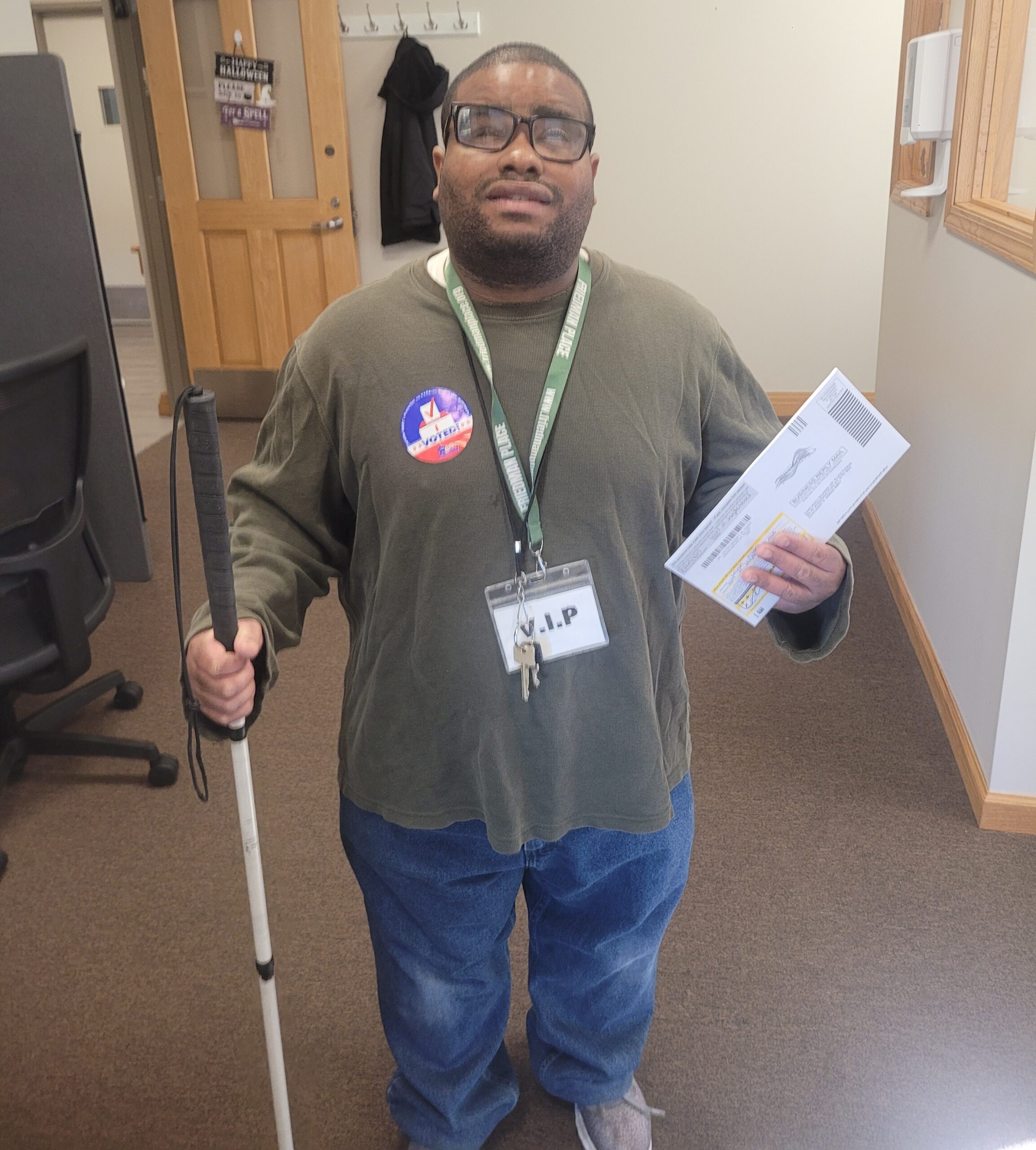 A smiling resident stands with their white cane and mail-in ballot with an "I voted" sticker.