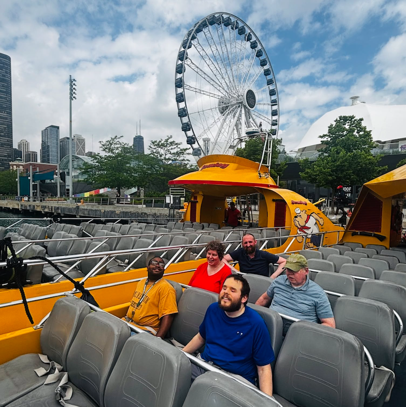 A group of residents sit on the top of a tour boat with the Navy Pier ferris wheel in the background.